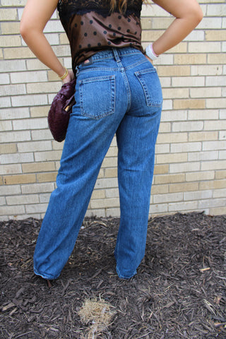 A person shown from the back wearing JBD Square Patch Pocket Straight Medium wash blue wide-leg jeans, a satin brown polka-dot top, and holding a dark purple clutch, available at Erin Rose Boutique