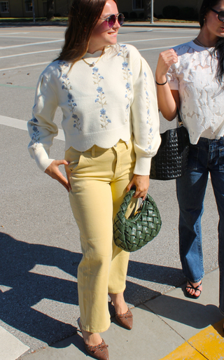 A woman walking outdoors on a sunny street wearing a cream knit sweater with blue floral embroidery, pale yellow cropped pants, and brown pointed flats, while holding a green woven handbag.