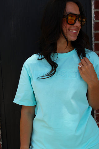 A woman wearing a light blue short-sleeve t-shirt with a subtle tonal logo on the chest, styled with dark sunglasses, standing against a dark wooden door and brick wall.