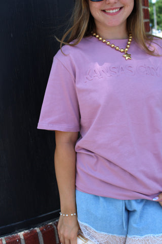Model wearing the KC Embossed Essential Tee in dusty lavender featuring tonal Kansas City lettering and a relaxed fit.