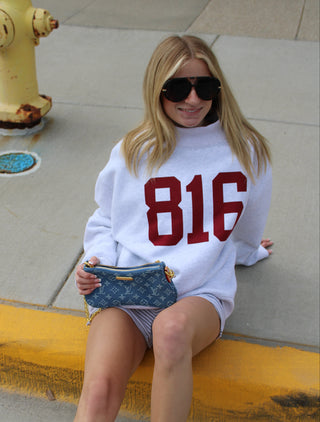 A woman sitting on a yellow curb wearing a white sweatshirt with '816' in red. 