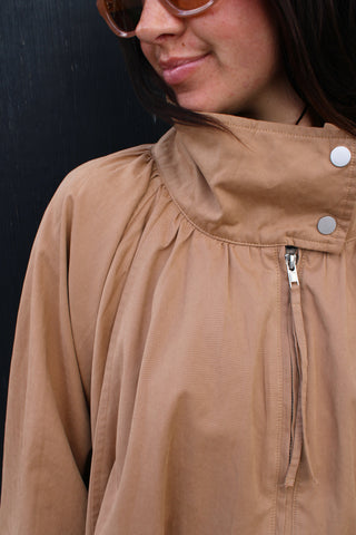 Close-up of a person wearing a tan high funnel neck jacket with a partial front zipper and two silver snap buttons near the collar, set against a dark background.