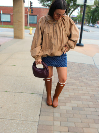 A woman stands on a city sidewalk wearing a Camel zip front jacket with high funnel collar with a blue patterned mini skirt, brown knee-high boots, and holding a dark purple handbag.