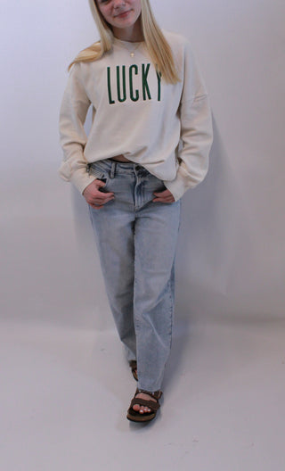 Person wearing a beige sweatshirt with 'LUCKY' text and light blue jeans on a white background