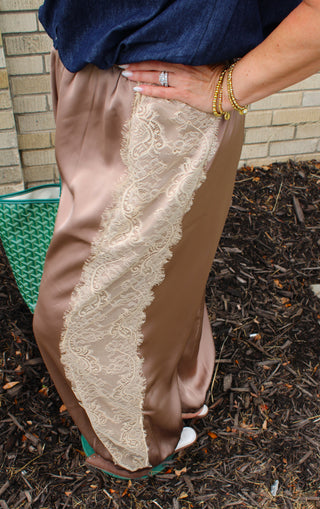 A close-up of a person wearing wide-leg Ivory lace side panel detail on mocha satin pants, paired with a navy top and gold bracelets available at Erin Rose Boutique
