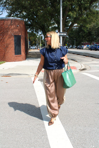 A woman walking across a street crosswalk wearing Women's mocha balloon pants styled with denim bubble top in an elevated casual outfit, and carrying a green woven tote bag.