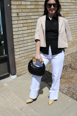 Woman in beige cropped blazer, black shirt, white pants, and sunglasses standing against a brick wall.