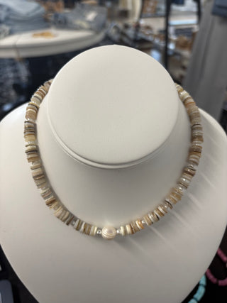 Neutral-toned puka shell necklace with natural disc beads and a center pearl accent, displayed on a white bust stand, beach-inspired jewelry