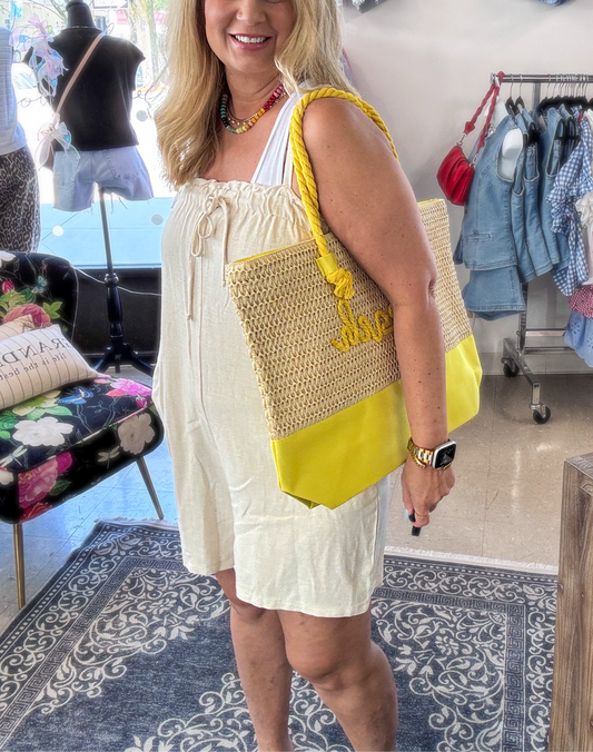 Woman wearing a light neutral romper with spaghetti straps, a smocked bodice, and drawstring neckline, styled with a yellow tote bag