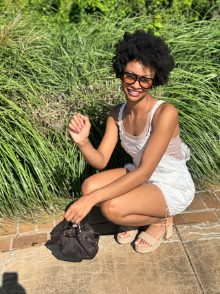 A woman wearing a white, lace  sleeveless romper, sunglasses, crouches outdoors on a stone patio in front of tall green ornamental grass, holding a chocolate brown textured handbag.