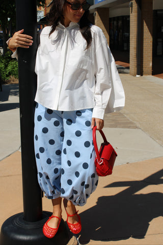 Woman wearing light blue polka dot balloon pants styled with a white poplin blouse and red flats, carrying a red handbag for a chic, elevated casual outfit