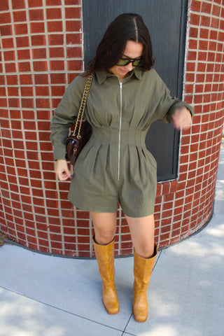 Woman wearing Pamela olive green long sleeve romper with pleated waist, front zipper and tan knee-high boots.