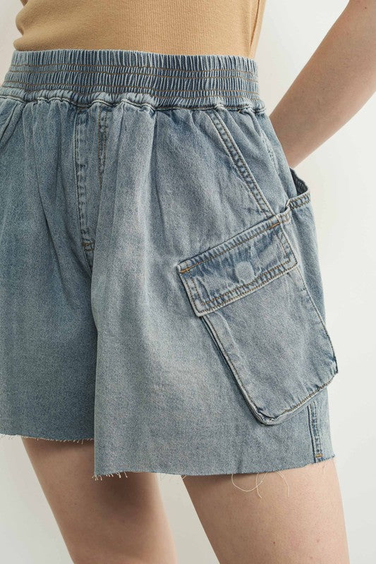Close-up of denim parachute shorts showing smocked waistband, utility pocket, and raw-edge hem