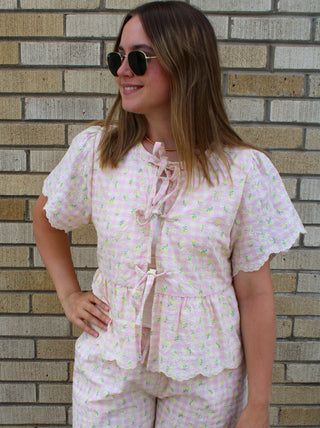 Woman wearing a pastel pink gingham blouse with yellow floral print and tie-front bow details, styled for a cute and feminine summer boutique look available at Erin Rose Boutique
