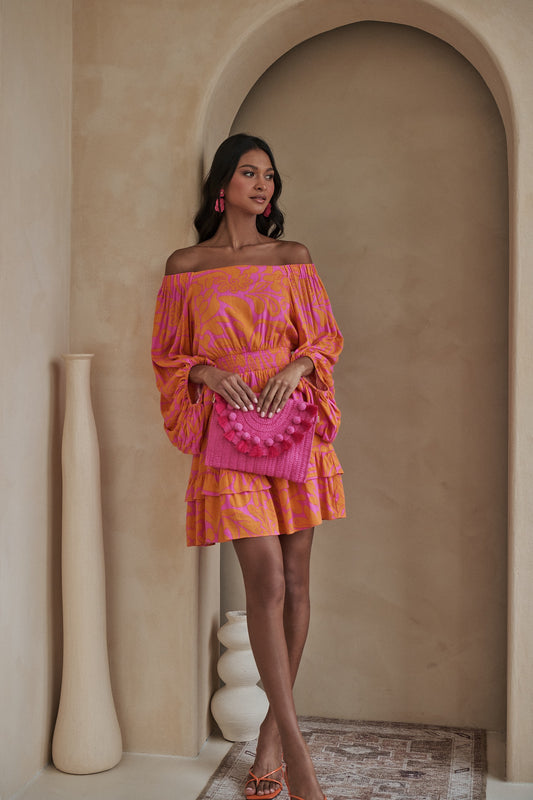 Woman wearing a pink and orange off-the-shoulder smocked mini dress with tiered ruffle hem now at Erin Rose Boutique.   