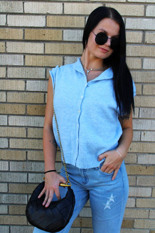 A woman wearing a light blue sleeveless knit piper cardigan vest shirt and light blue jeans with star patches, holding a black woven crossbody bag, standing against a beige brick wall.