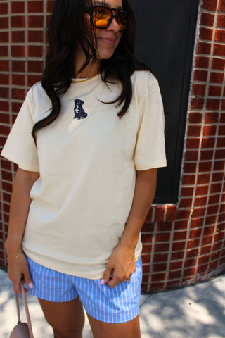 Model wearing the Pup Club Embroidered Tee in buttercream featuring a small embroidered puppy graphic, styled with blue striped boxer shorts for a casual summer outfit.