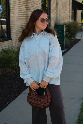 A woman wearing a light denim blue and heather gray striped long-sleeve collared rugby shirt, dark brown pants, and sunglasses, holding a brown woven handbag on a sidewalk next to a brick building.