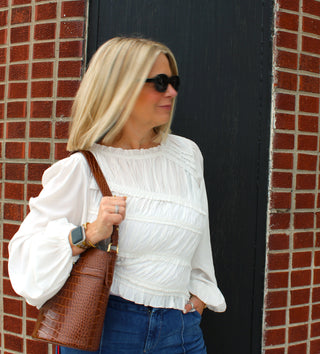 A woman with blonde hair wearing a white long-sleeve , ruched, asymmetrical, ruffled blouse with blue jeans, and black sunglasses, carrying a brown crocodile-embossed leather shoulder bag. She is standing in front of a black door framed by red brick walls.