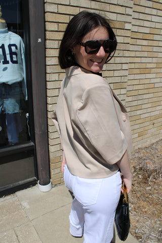 Woman wearing sunglasses and a beige jacket standing outside a building with a brick wall.