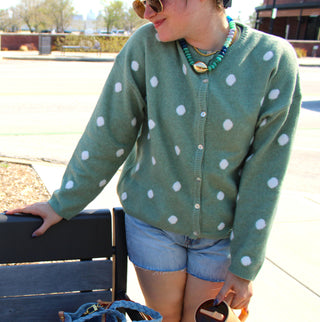 Person wearing a sage green polka dot cardigan sweater and denim shorts outdoors.