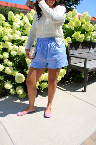 Model wearing Sail Away Striped Boxer Shorts featuring blue and white pinstripes, an elastic waistband, faux button detail, and side pockets styled with a white knit sweater.