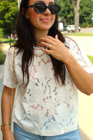 A woman wearing a white floral lace short-sleeve top, blue jeans, and black sunglasses.