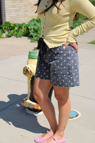 A person outdoors wearing a pale yellow ribbed long-sleeve top and dark gray denim shorts with white polka dots. The person is standing on a concrete sidewalk next to a yellow fire hydrant.
