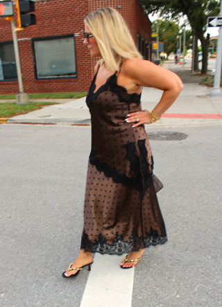 A woman wearing a sheer brown polka-dot maxi skirt and matching cami with black lace trim stands on an urban street corner.