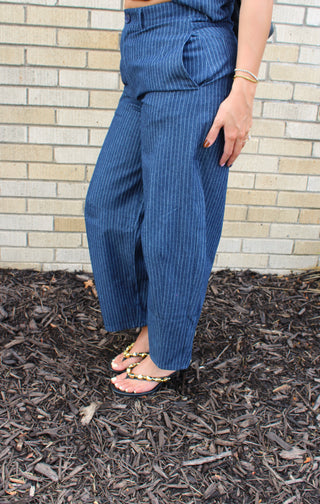 A person wearing blue pinstriped wide-leg pants and heeled sandals, standing outdoors in front of a light beige brick wall.