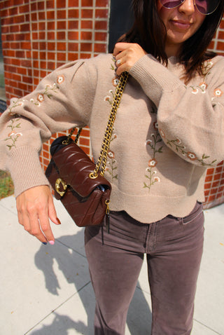 A woman wearing a taupe knit sweater with delicate floral embroidery, holding a dark brown leather shoulder bag with a gold chain strap. She is wearing brown pants and sunglasses.