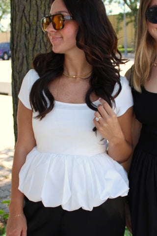 Women's white bubble hem peplum top with a square neckline and short sleeves, styled with black pants and gold jewelry.