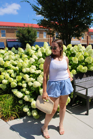 Model wearing a white open knit sleeveless sweater tank styled with blue gingham bubble shorts and neutral heels.