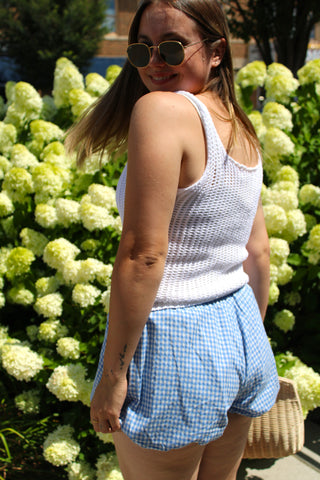 Model wearing a white open knit sleeveless sweater tank styled with blue gingham bubble shorts and neutral heels.