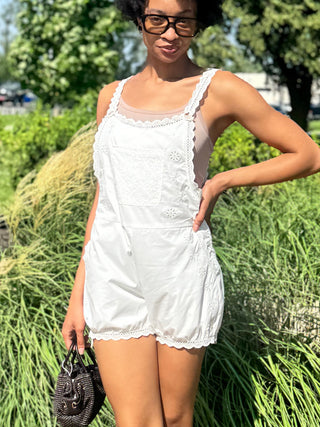 A woman wearing a white sleeveless overall romper with lace trim stands outdoors and holding a small dark handbag.