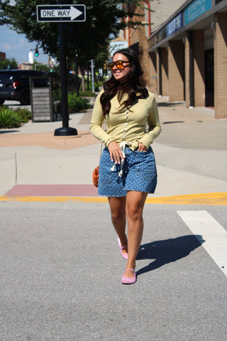 A woman walking on a city street wearing a pale yellow long-sleeve button-up top, a blue denim mini skirt, pink flat shoes, and orange-tinted sunglasses.