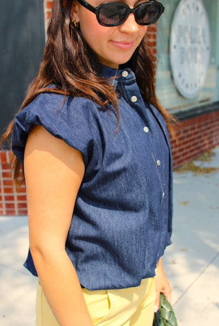 A woman wearing a dark blue chambray short-sleeve button-up Women's boutique denim top with bubble hem and yellow pants, standing outdoors on a sidewalk in front of a brick building.