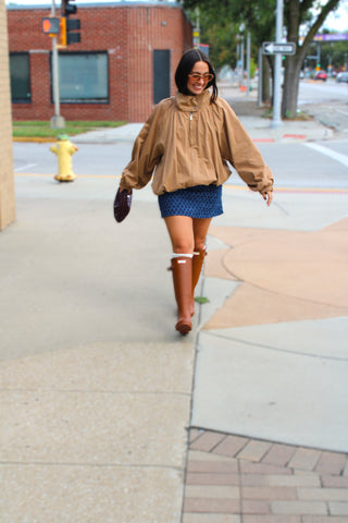 A woman walking on a city sidewalk wearing a loose tan oversized jacket with bubbled hem & funnel neck collar, a blue patterned mini skirt, and tall camel brown hunter boots.