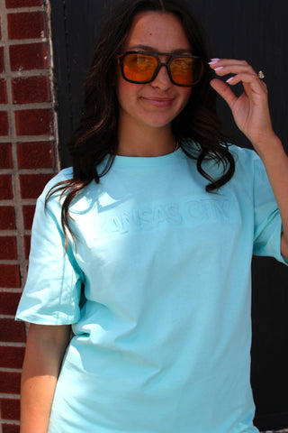 A woman wearing a light aqua blue short-sleeve t-shirt with 'KANSAS CITY' printed in tonal letters across the chest, paired with orange-tinted sunglasses, standing outdoors against a dark door and brick wall.