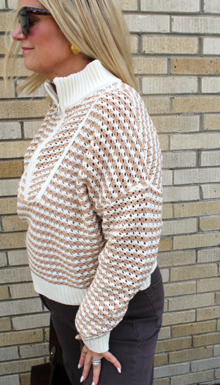 A woman wearing a cream and brown waffle-knit mock neck sweater, standing in front of a beige brick wall.