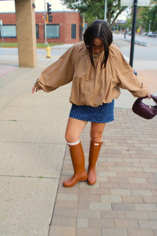 A woman stands on a city sidewalk wearing the oversized Ken Funnel Neck Jacket styled with denim patterned mini skirt, and tall tan rain boots, while holding a dark purple handbag.