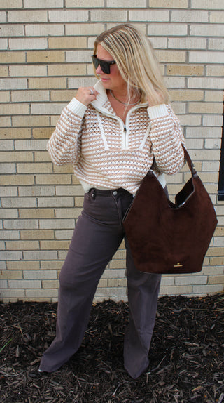 A woman wearing a cream and brown patterned preppy knit half-zip pullover, dark brown flared jeans, and sunglasses, while carrying a large brown suede shoulder bag.