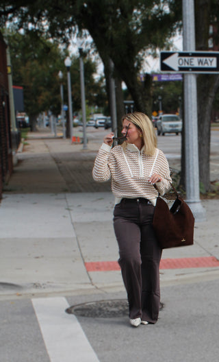 A woman walking on a city sidewalk wearing a beige and white striped half zip long-sleeve sweater, dark brown wide-leg dear john pants, and carrying a large brown tote bag.