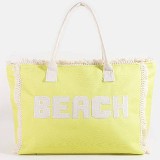 Bright yellow canvas beach tote bag with white “BEACH” lettering and fringe trim, featuring sturdy white handles and a roomy interior.