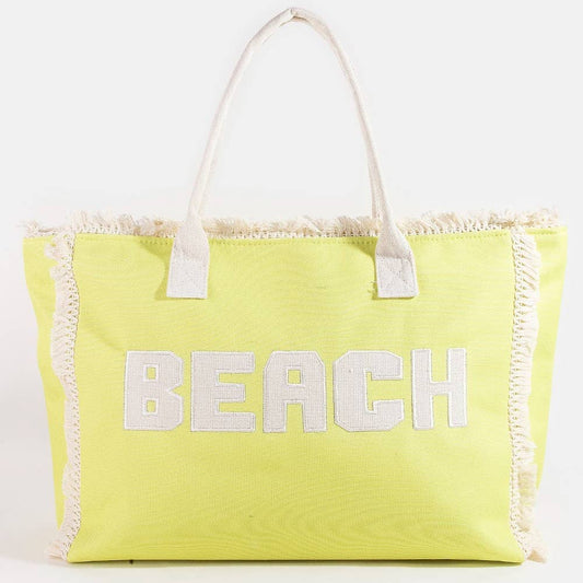 Bright yellow canvas beach tote bag with white “BEACH” lettering and fringe trim, featuring sturdy white handles and a roomy interior.