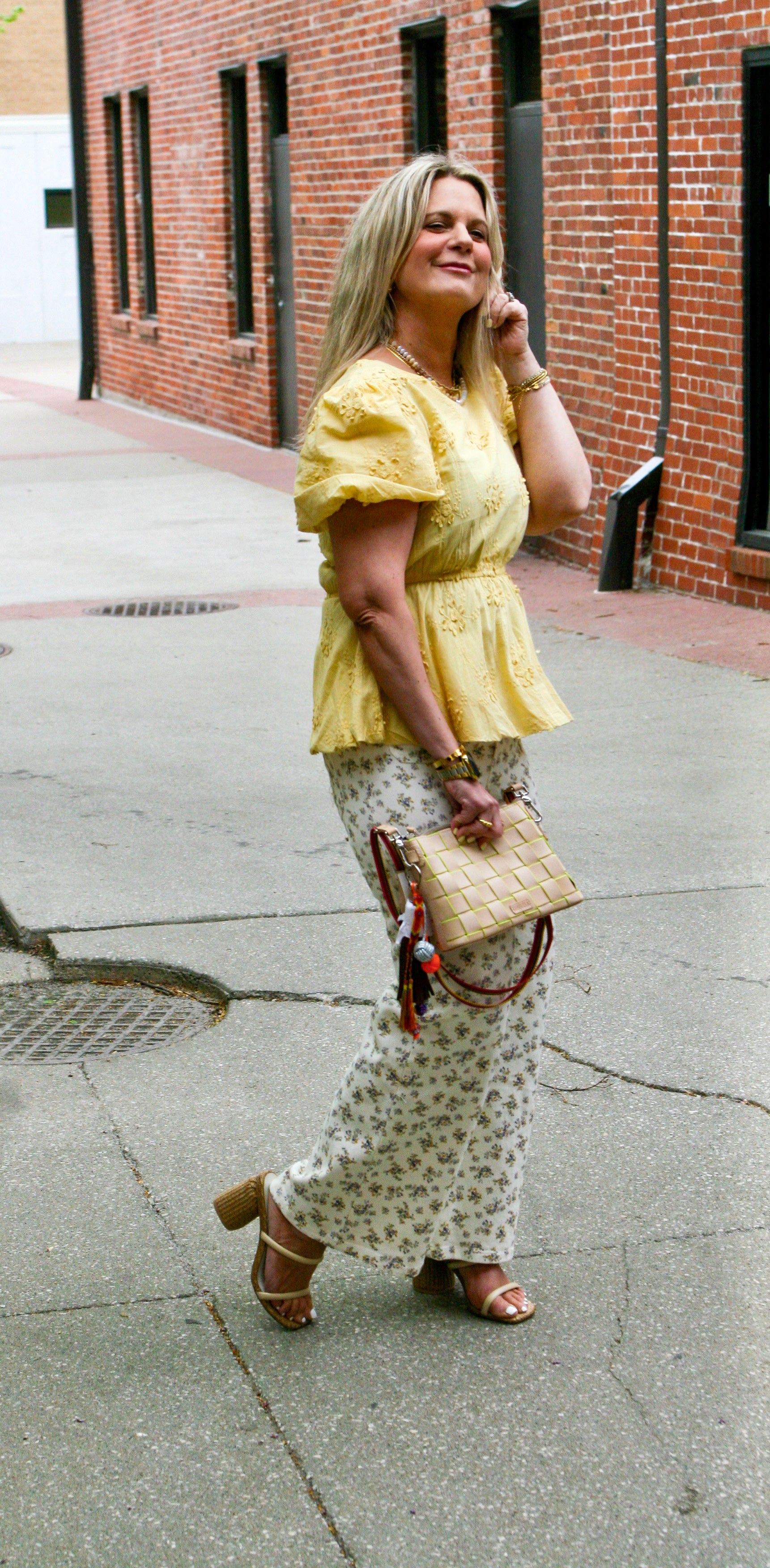 Model wearing yellow ruffle sleeve embroidered top with white floral pants and woven clutch handbag, smiling and walking outside – Erin Rose Boutique